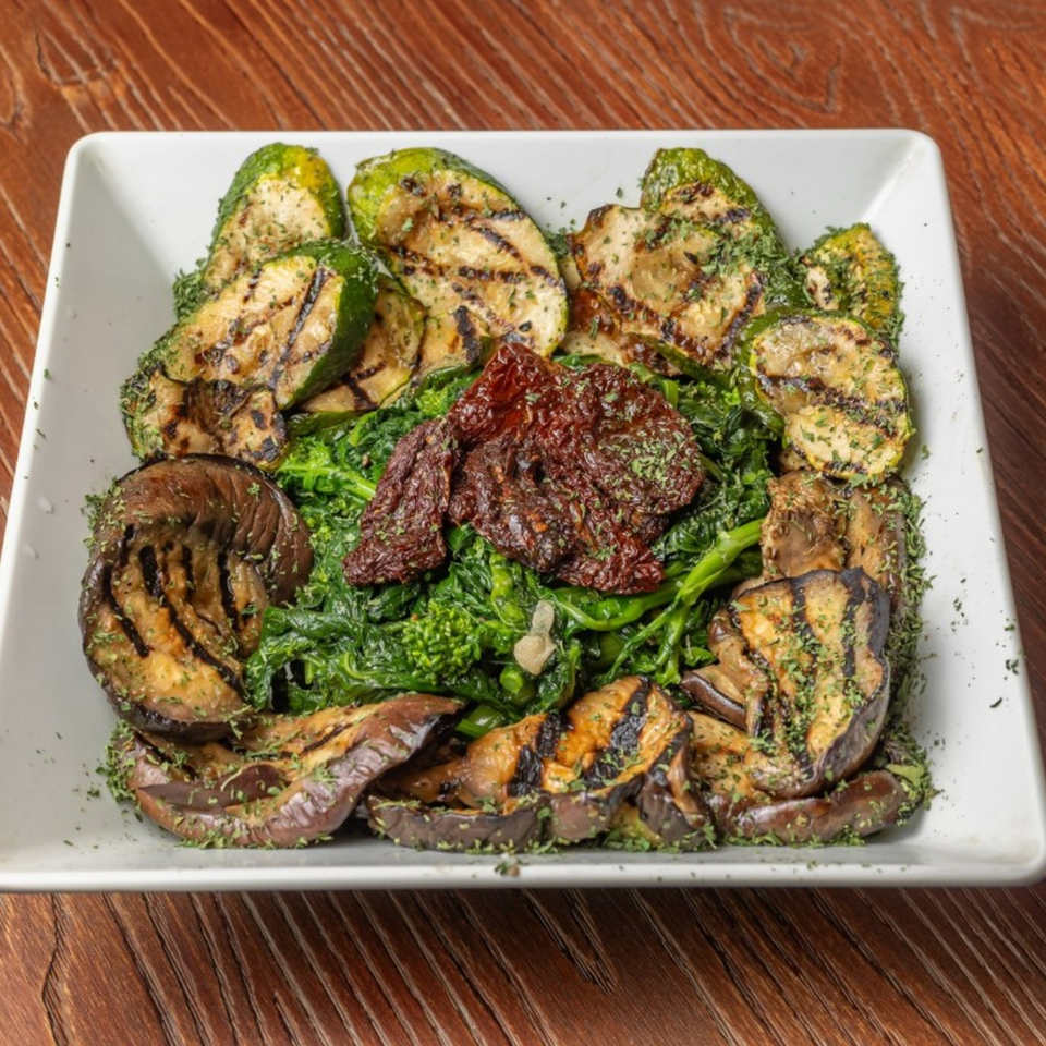 Grilled zucchini and eggplant with broccoli rabe and sun-dried tomatoes on a white plate.